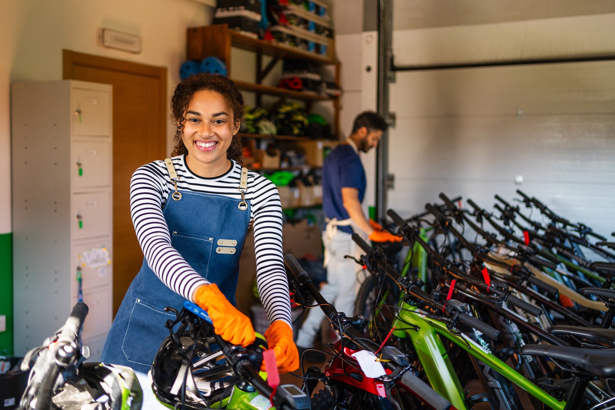 Woman in apron and gloves preparing an e-bike for rental in a bright service garage, smiling and confident while working